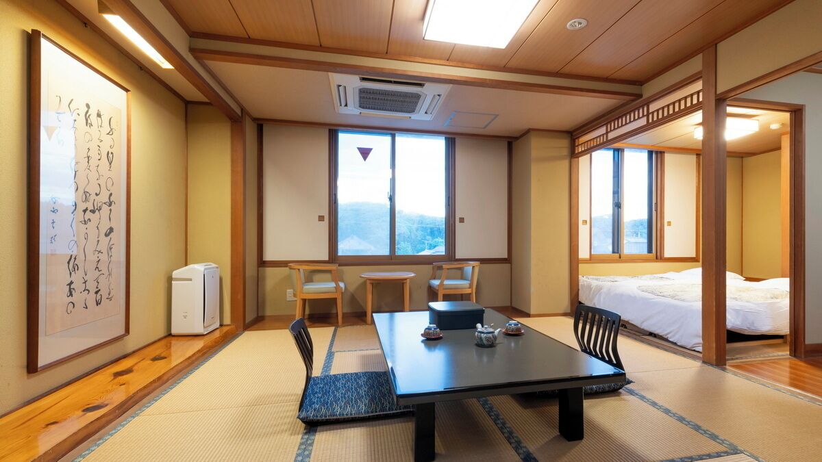 A traditional Japanese room with tatami flooring, a low table with a tea set, a bed with a white bedspread, and a large window with a view.
