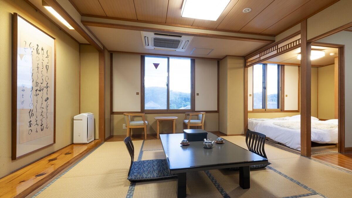 Una habitación japonesa tradicional con suelo de tatami, una mesa baja con vajilla de té, una cama con edredón blanco y un ventanal con vistas.