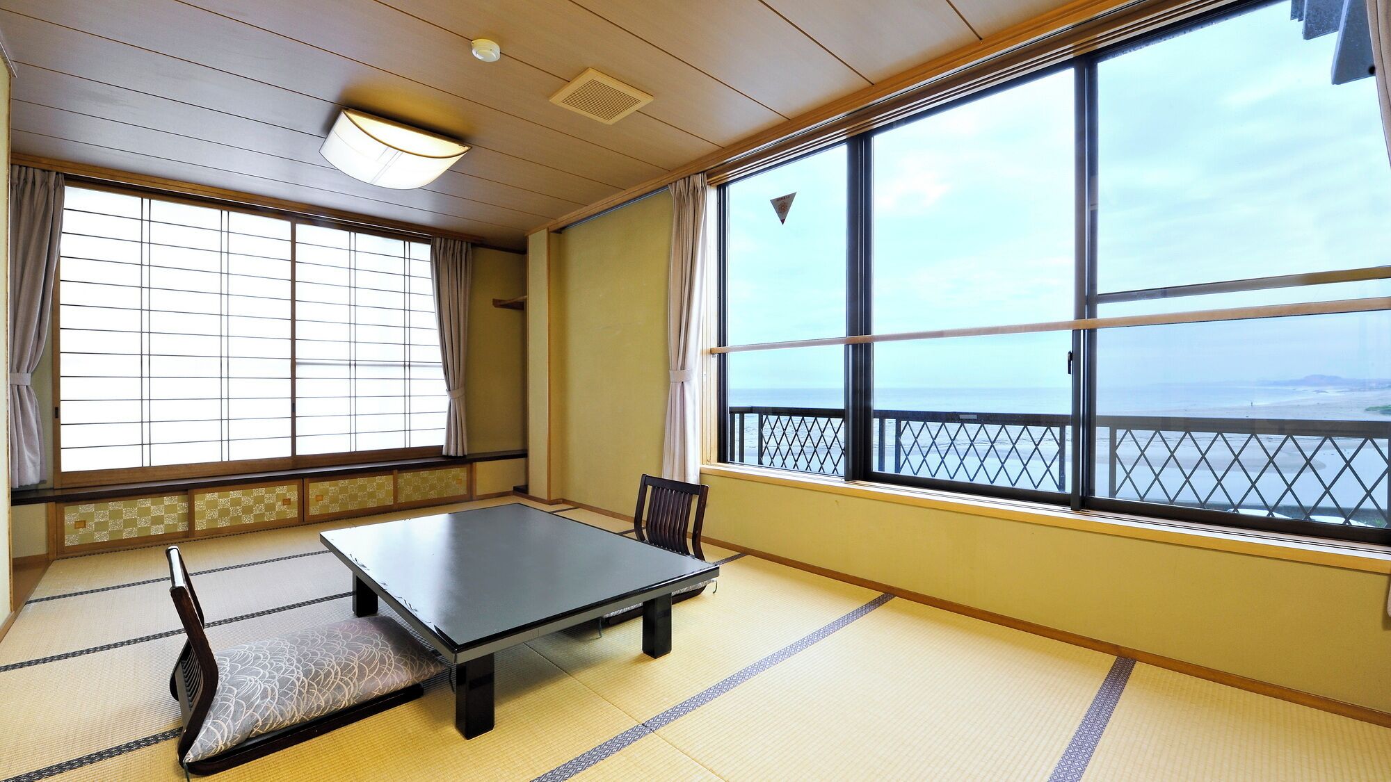 Una habitación con tatami, una mesa baja y una silla, ventanas amplias con vistas al mar y puertas correderas tradicionales.