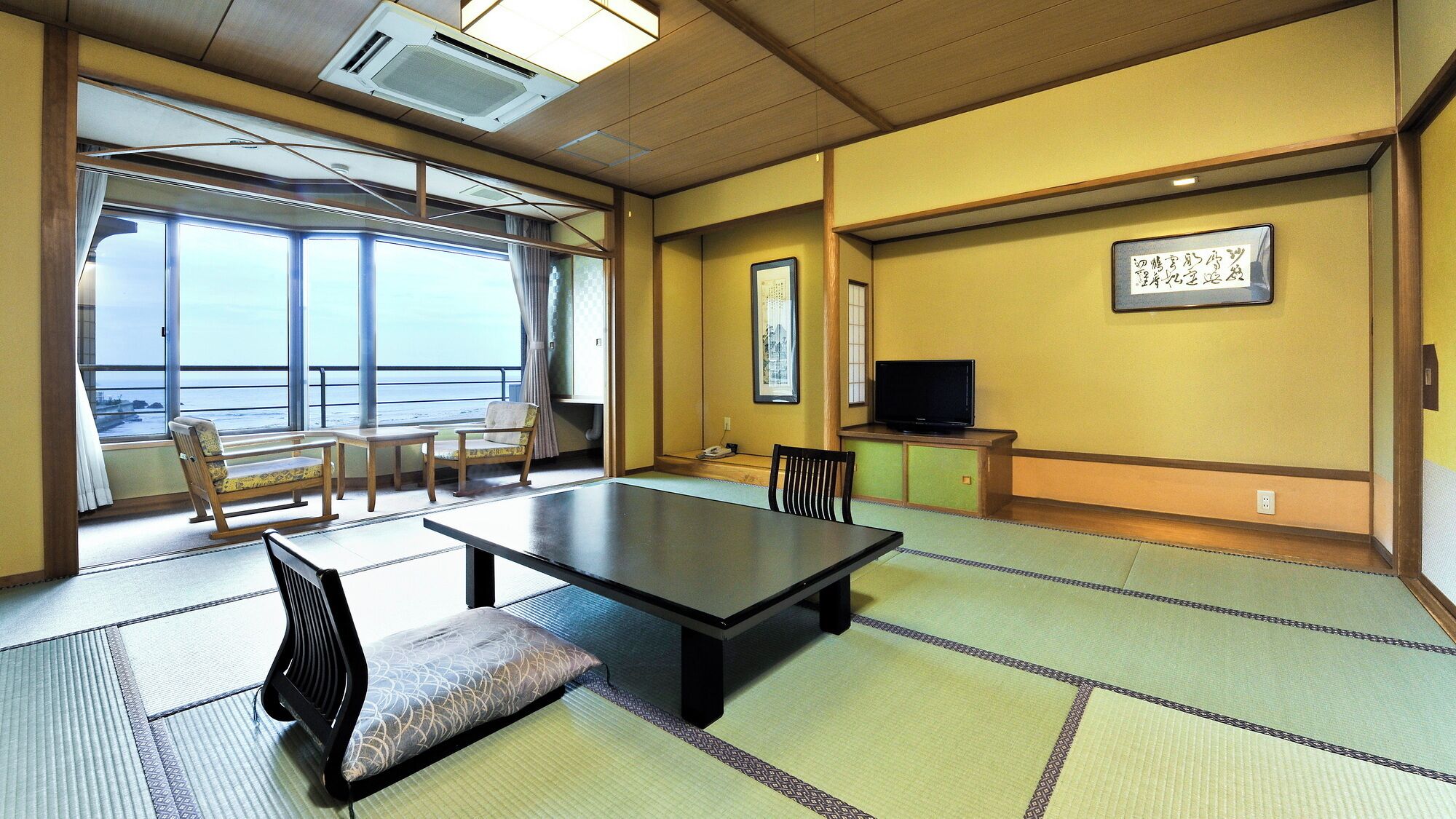 A room with tatami flooring, a low table, chairs, a television, and a window with a view.