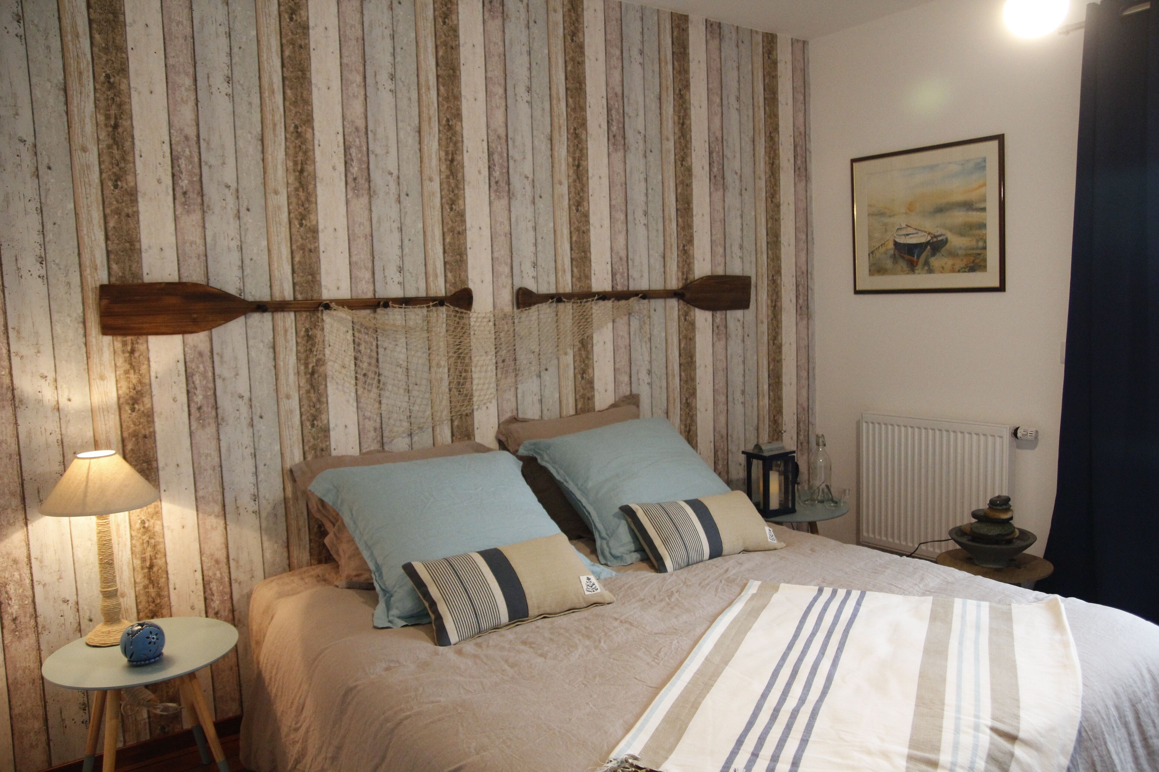 A bedroom with a wooden slat wall, a bed with striped bedding, a small bedside table with a lamp, and a framed picture on the wall.