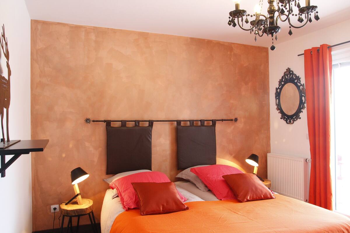 A bedroom with a bed, two nightstands, a chandelier, a radiator, and a window with curtains.