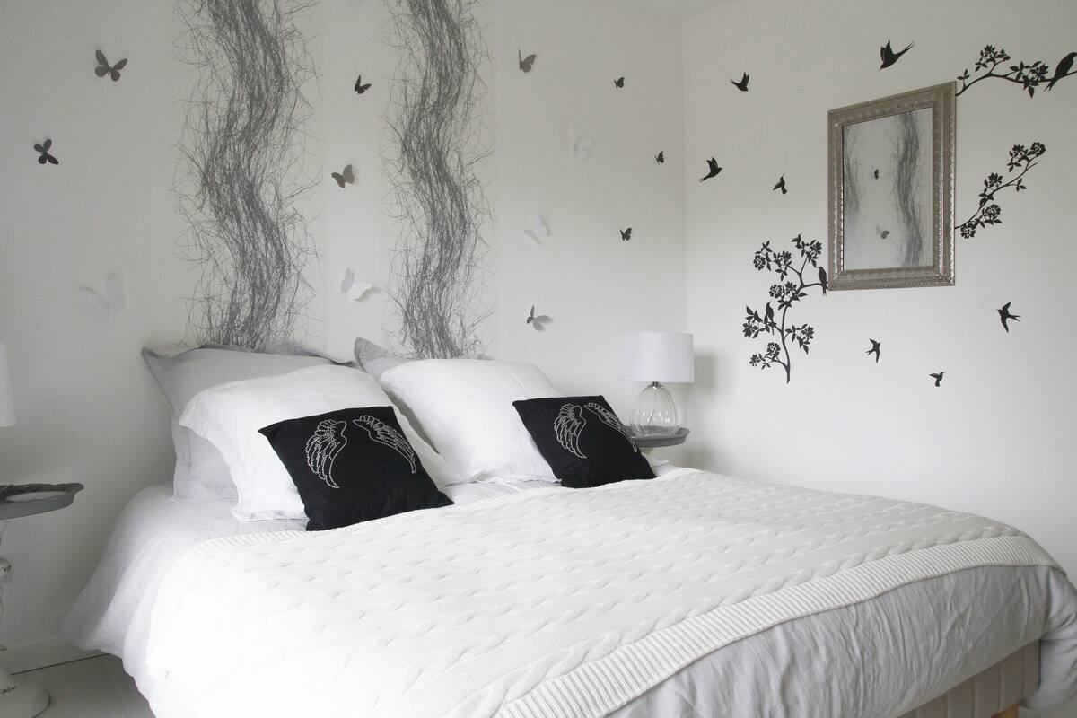 A neatly made bed with white bedding and two black pillows with wing designs, a bedside table with a lamp, and wall decorations including butterflies and a framed picture.