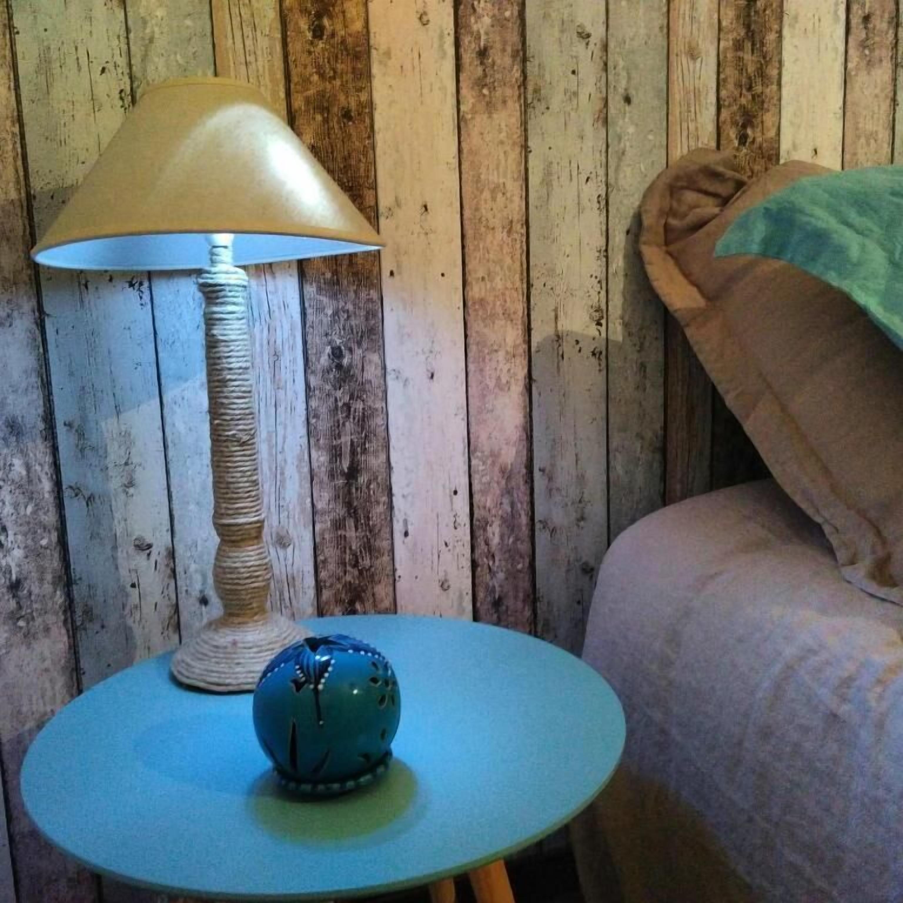 A room with a round table, a lamp, and a bed against a wooden wall.