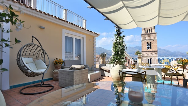 A terrace with outdoor furniture, a hanging chair, a table with a glass top, and a view of a tower and mountains. - B&B La Gaetana (Gaeta)