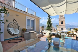A terrace with outdoor furniture, a hanging chair, a table with a glass top, and a view of a tower and mountains. - B&B La Gaetana (Gaeta)