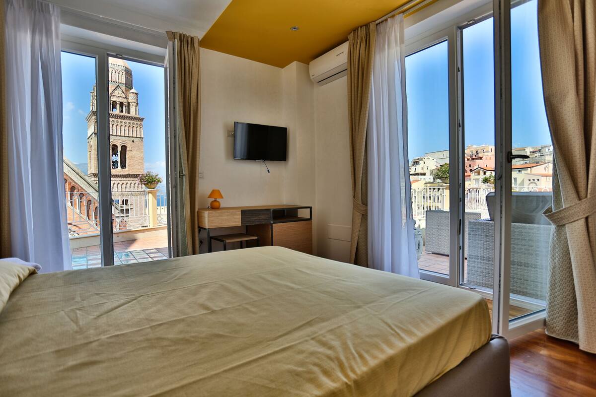 A hotel room with a large bed, a desk, a TV, and a balcony with a city view.