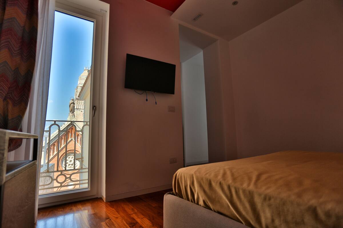 A bedroom with a bed, a TV mounted on the wall, and a balcony with a view of a building.