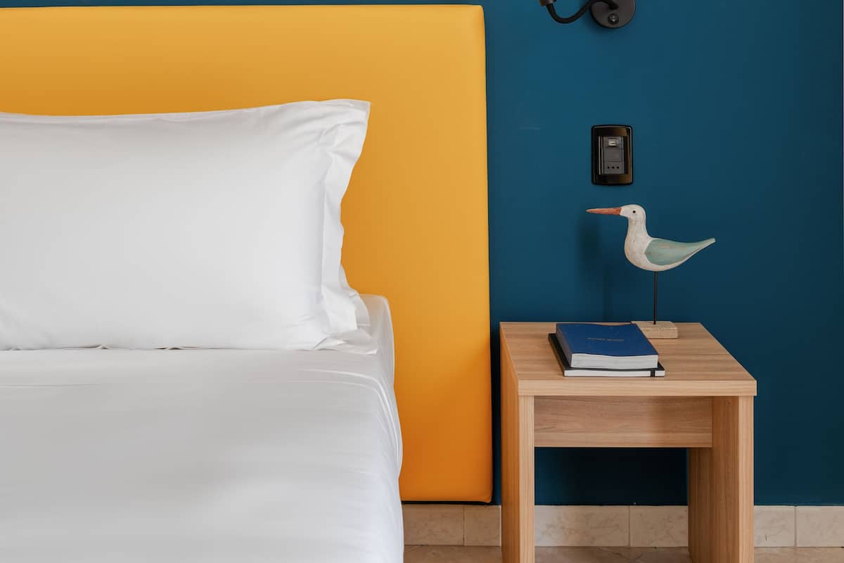 Una cama con sábanas blancas, una mesita de noche de madera con un libro y una lámpara, y una pared pintada de azul y naranja.