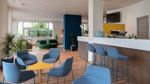 Uma área de bar moderna com cadeiras azuis e uma mesa amarela, uma cozinha com televisão e um sofá verde numa sala clara com janelas grandes.