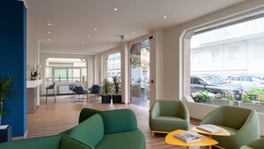 Um lobby moderno com assentos verdes, piso de madeira e grandes janelas com vista para a rua.