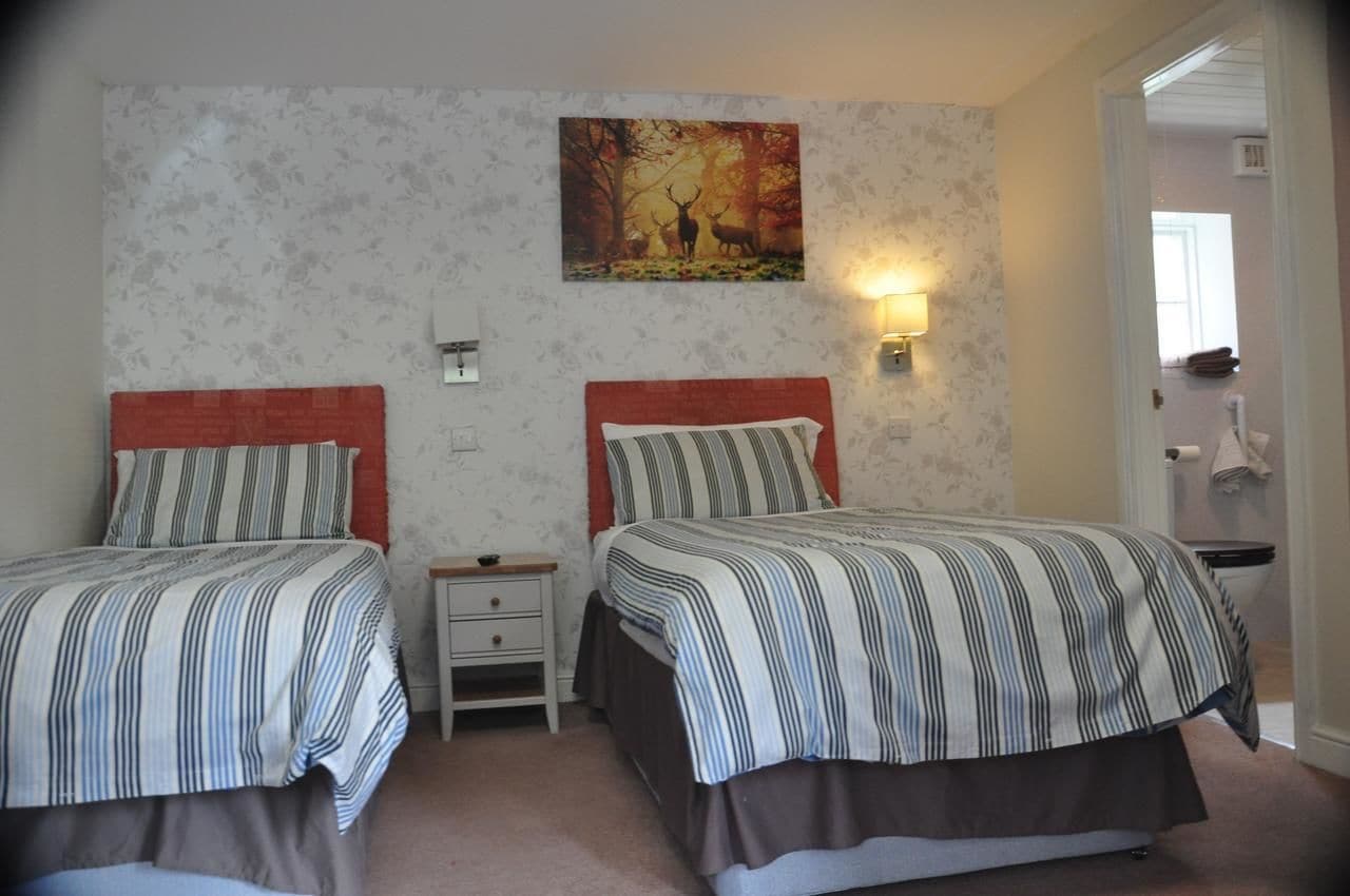 Une chambre avec deux lits, une table de chevet, une lampe fixée au mur et un tableau représentant des cerfs dans une forêt.