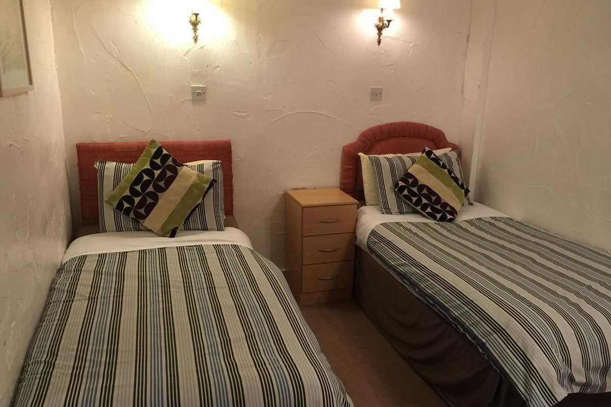 A room with two single beds, each with striped bedding and patterned pillows. There is a small wooden nightstand between the beds.