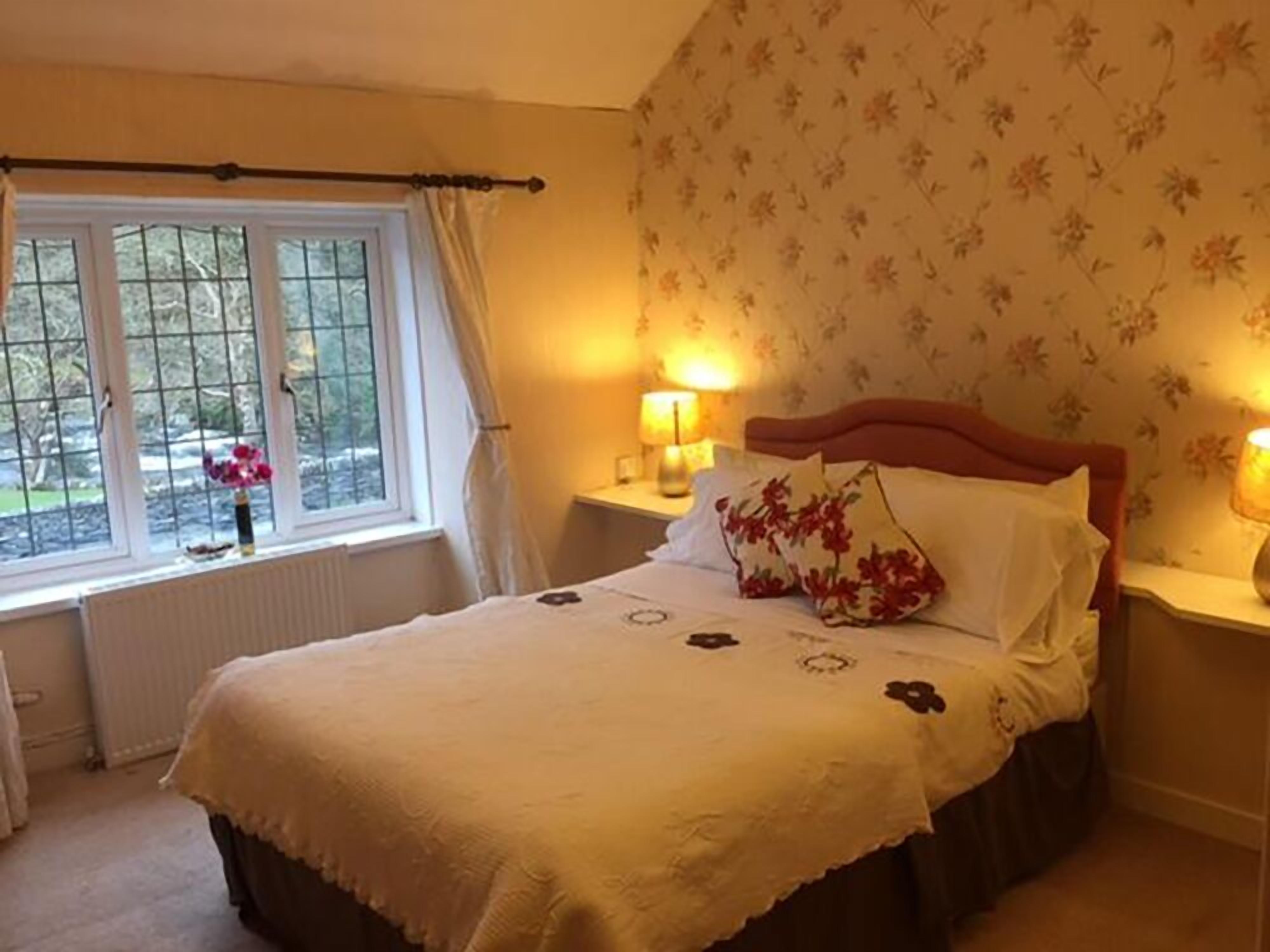 A bedroom with a bed, two bedside tables with lamps, a window with curtains, and floral wallpaper.
