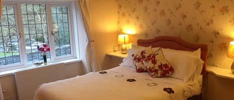 A bedroom with a bed, two bedside tables with lamps, a window with curtains, and floral wallpaper.