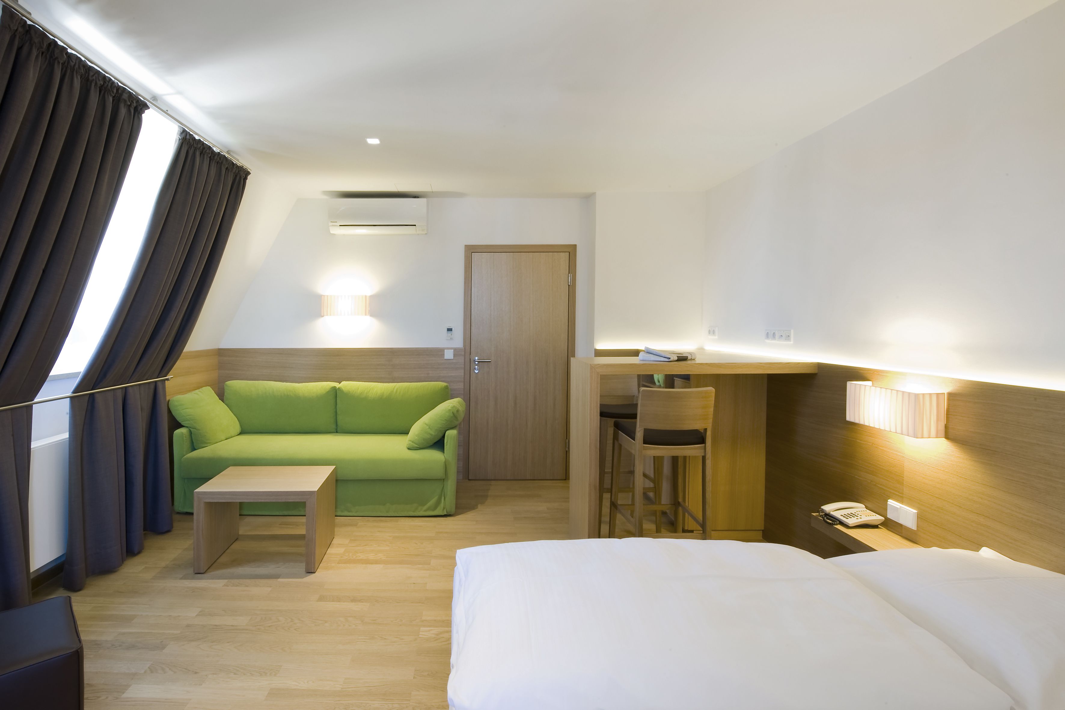 A hotel room with a bed, a green sofa, a small table, a desk with a chair, a phone, and a door.