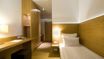 Quarto de hotel com uma escrivaninha de madeira, uma cama com roupa de cama branca e uma mesa de cabeceira com abajur.