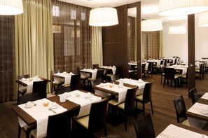 A hotel dining area with multiple tables set for dining, large windows with curtains, and modern lighting fixtures. - Hotel Thalmair (Munich)