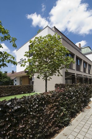 A modern hotel with a flat roof, large windows, and a well-maintained garden with a tree and hedges. - Hotel Thalmair (Munich)