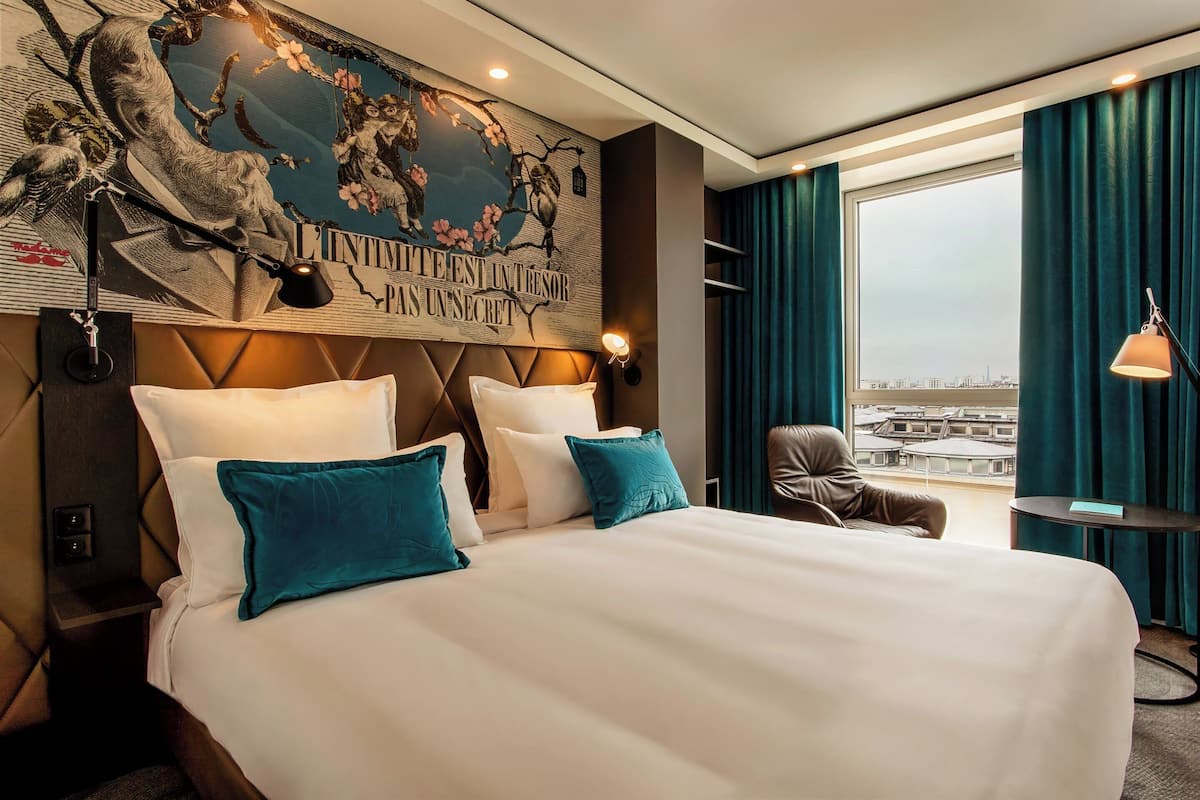 A hotel room with a large bed, teal pillows, a city view, a wall mural, and a small table with a lamp.