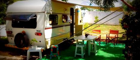 A white RV parked on a green surface with a table and chairs, a small stool, and a potted plant.