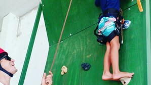 A child climbing a green climbing wall with various holds and a red triangular hold at the top. - Hostel Clube da Aventura (Conde)