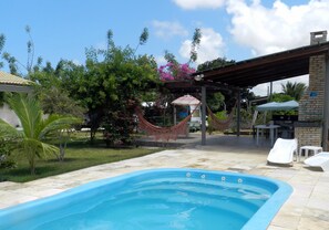 Una piscina con pérgola y hamaca, rodeada de follaje y un edificio de ladrillo.