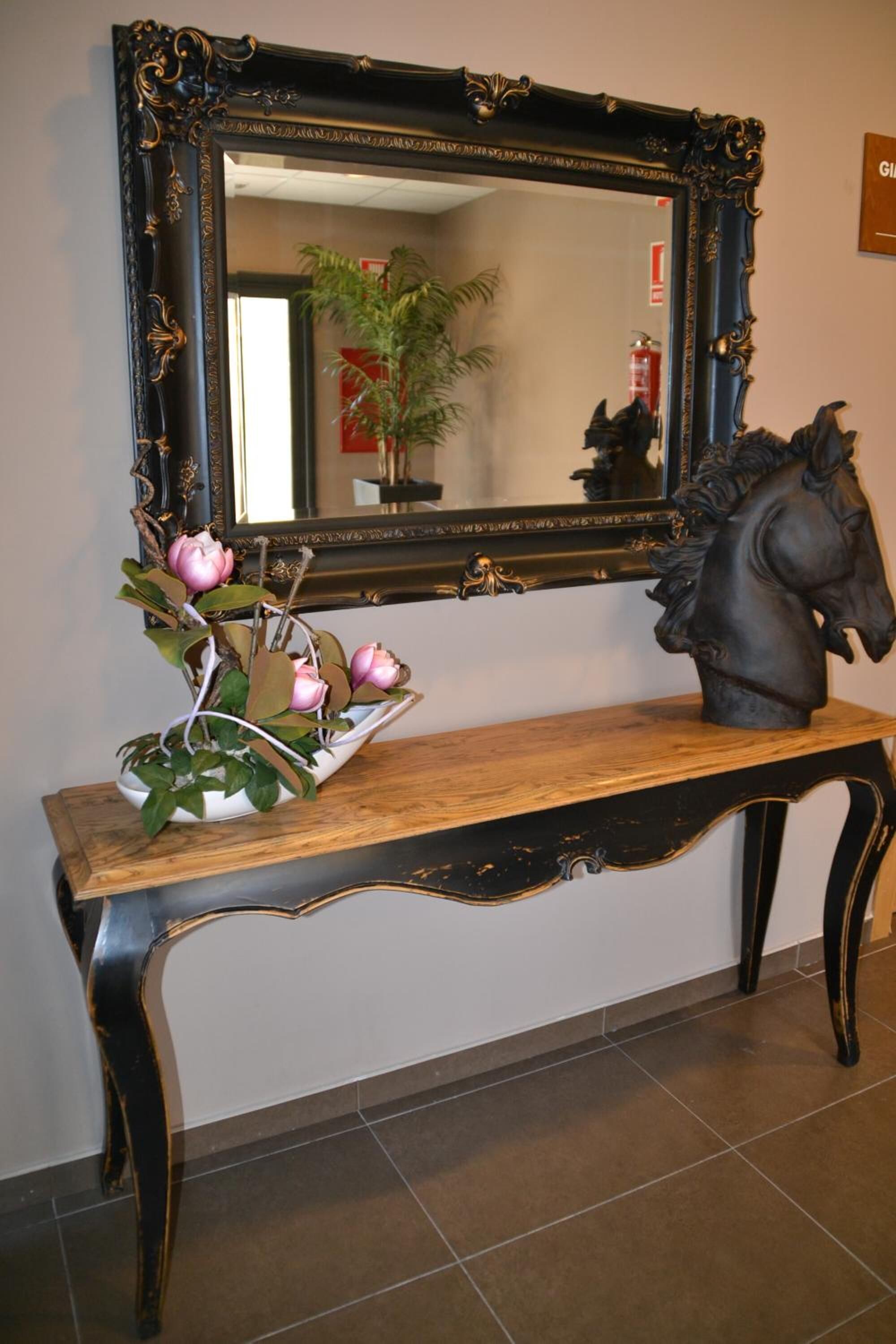 Una consola de madera con un espejo negro decorativo encima, una escultura de un caballo ornamental y un jarrón con flores rosadas.