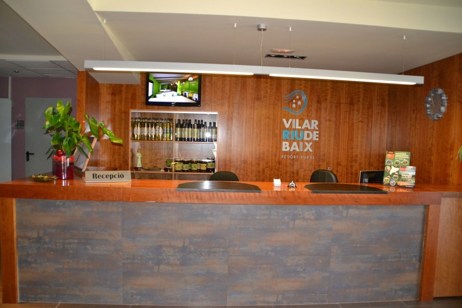 Una recepción con detalles de madera, un televisor montado encima y una pared con un reloj y un cartel que dice "Vilar Ruiz Baix Resort Rural".
