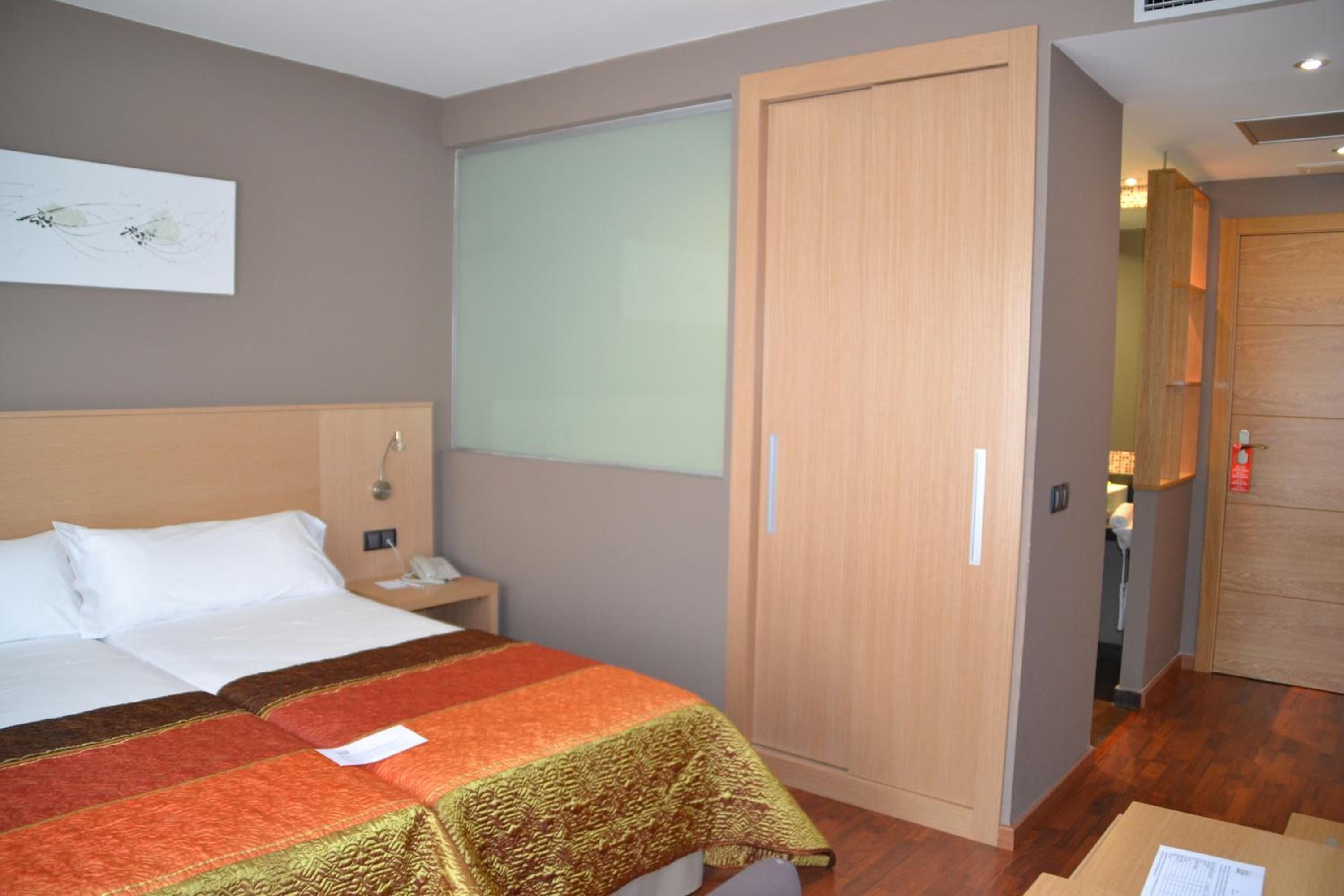A hotel room with a bed, a nightstand, a wooden wardrobe, and a window with blinds.