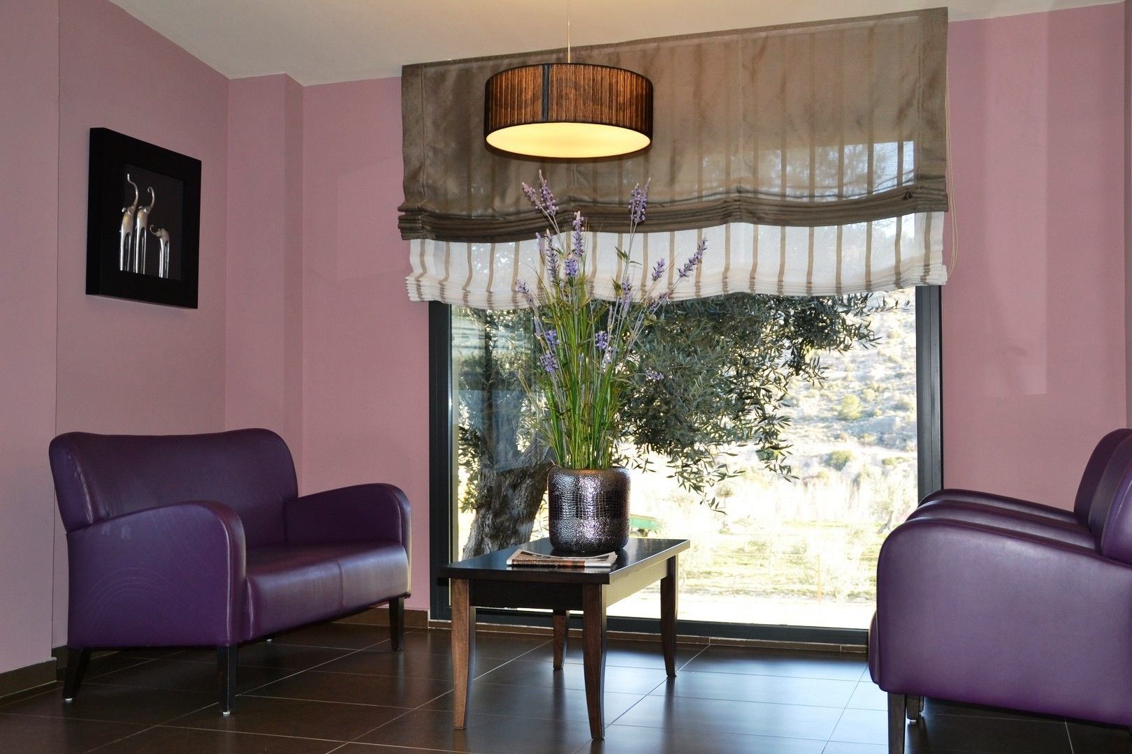 A room with two purple chairs, a table with a vase of flowers, a large window with a view of trees, and a wall-mounted lamp.