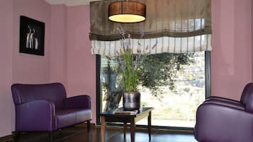 A room with two purple chairs, a table with a vase of flowers, a large window with a view of trees, and a wall-mounted lamp.