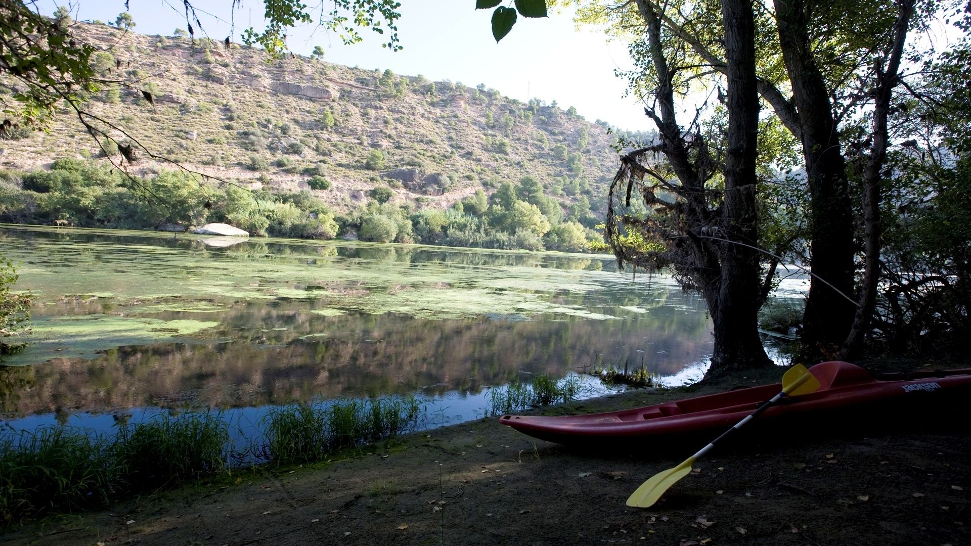 A calm river with a red canoe and paddle, surrounded by trees and a hilly landscape.