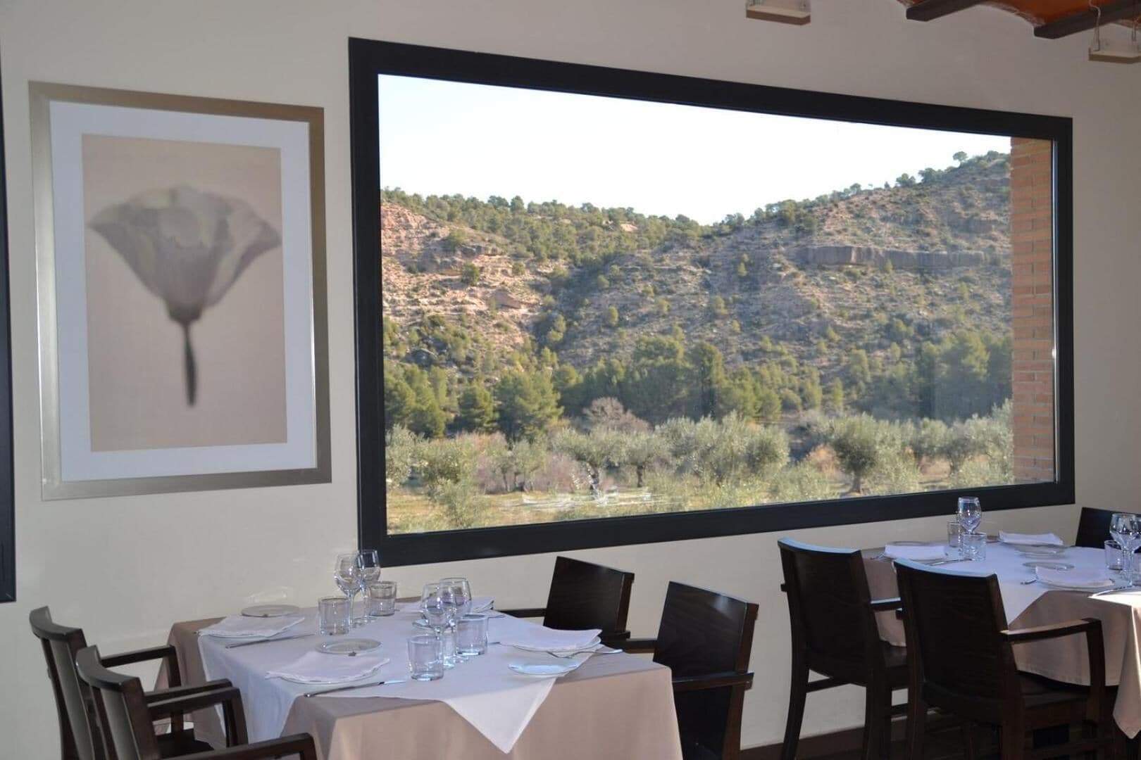 A dining area with tables set for four, a large window with a view of a mountainous landscape, and a framed artwork on the wall.