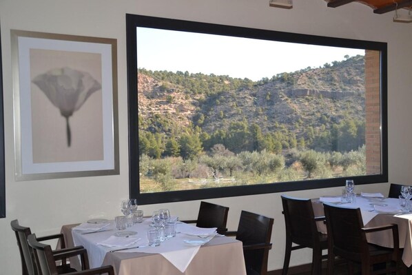 A dining area with tables set for four, a large window with a view of a mountainous landscape, and a framed artwork on the wall.