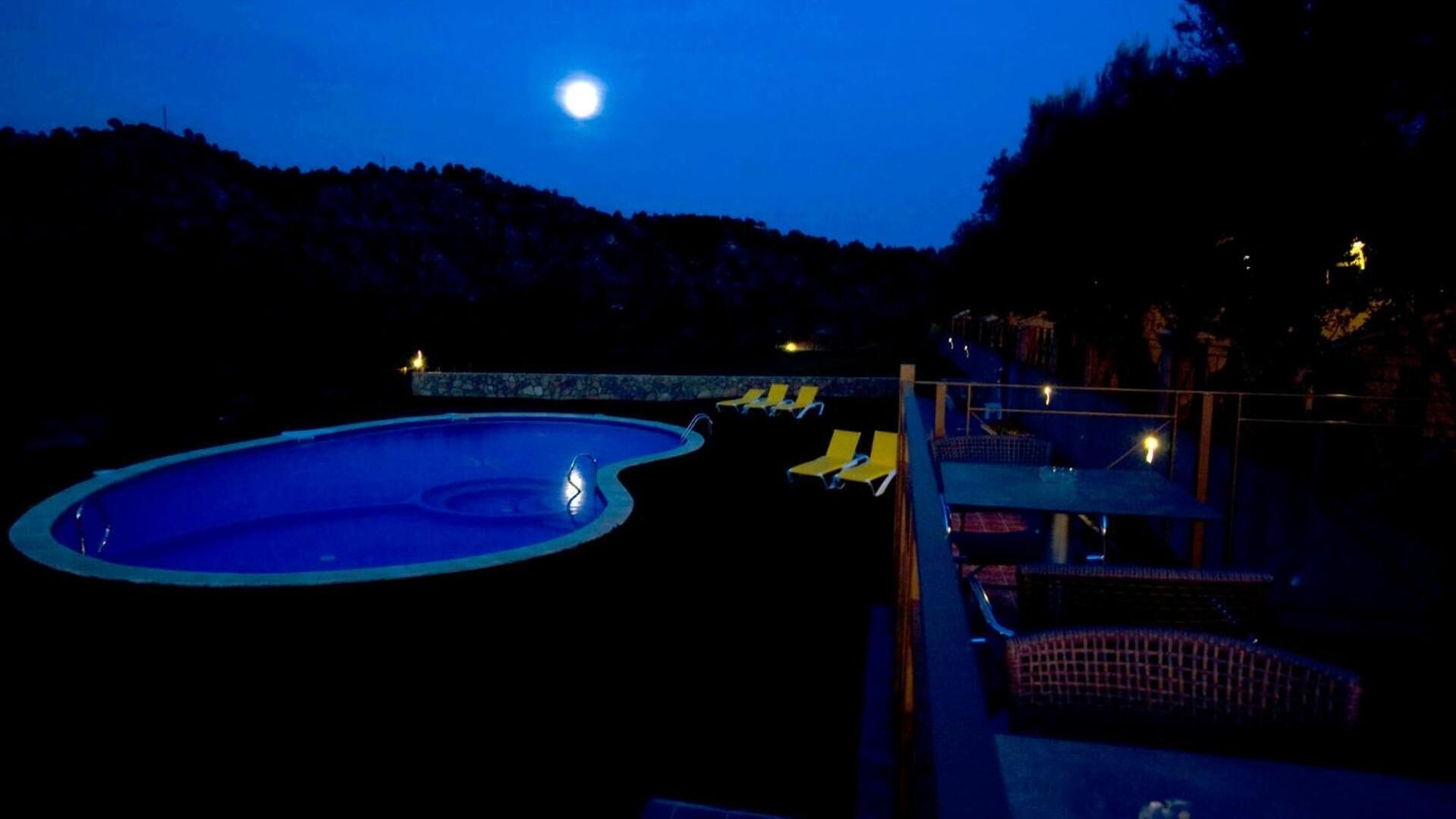 A swimming pool with a moon in the background, surrounded by lounge chairs and a mountainous landscape.