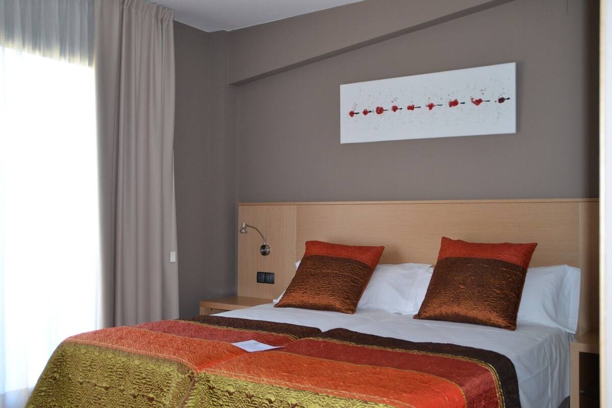 A hotel room with a bed, a nightstand, a wall-mounted lamp, and a window with curtains.