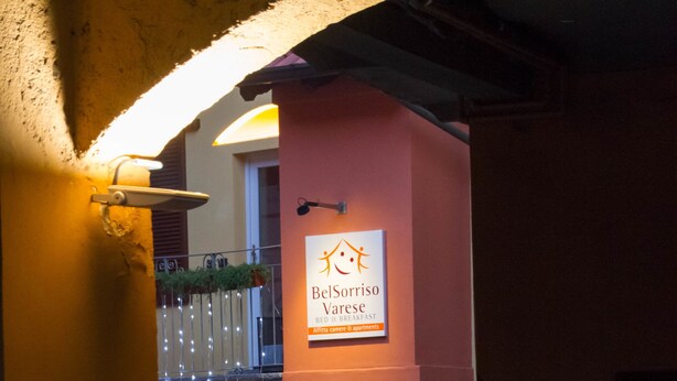 An entrance to a building with a sign for 'BelSorriso Varese' indicating a bed and breakfast. There are potted plants and a balcony with hanging lights. - Bel Sorriso Varese - Dormire Felice Rooms & Apartments (Varese)
