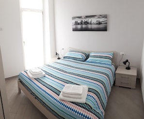 A modern bedroom with a large bed, a nightstand, a lamp, and a cityscape painting on the wall.