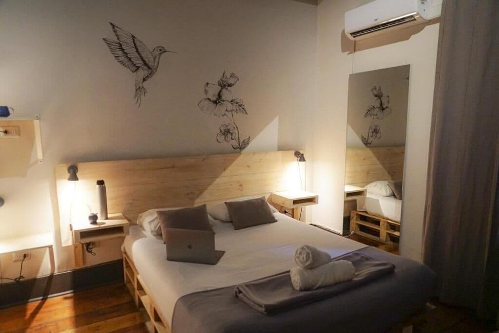 A bedroom with a wooden headboard, a bed with pillows, a laptop, a bedside lamp, and wall art of a bird and flowers.