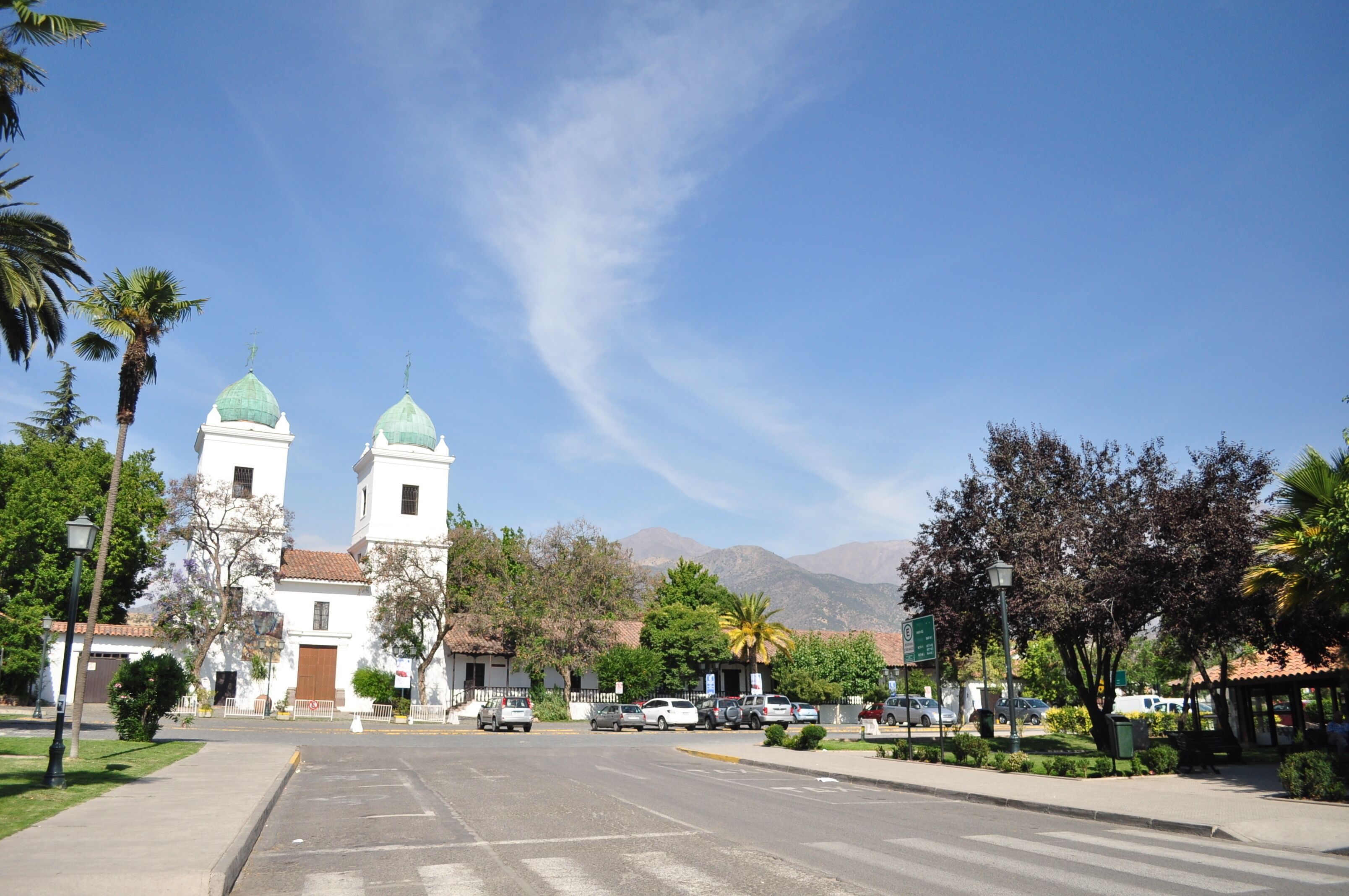 A street with a white building with two green domes, parked cars, and palm trees.
