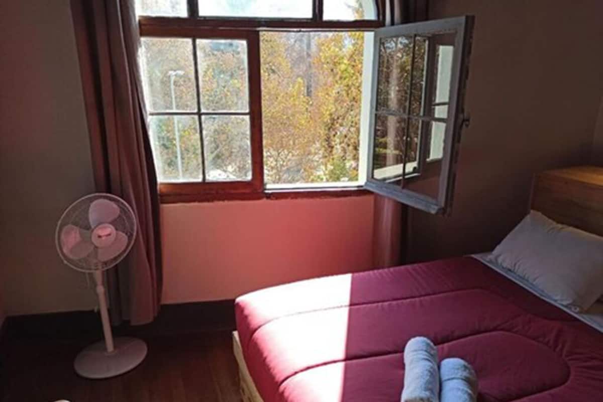 A bedroom with a bed, a window, a standing fan, and a wooden floor.