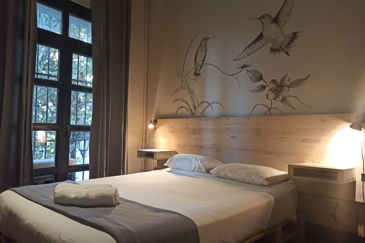 A bedroom with a wooden headboard, a bed with white linens, a bedside table with a lamp, and a window with curtains.