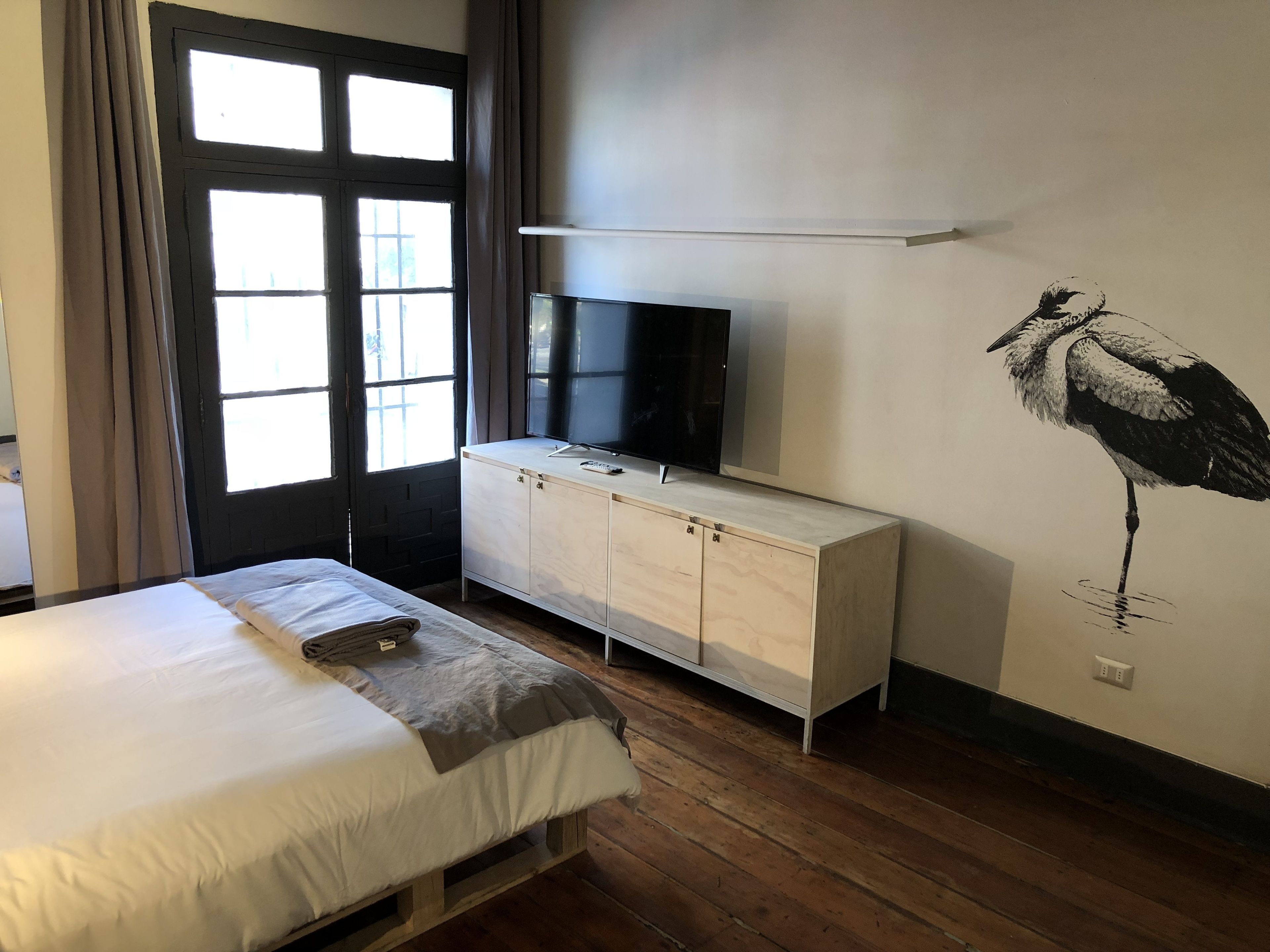Un dormitorio con cama, televisión sobre un mueble de madera y un mural de un pájaro en la pared.