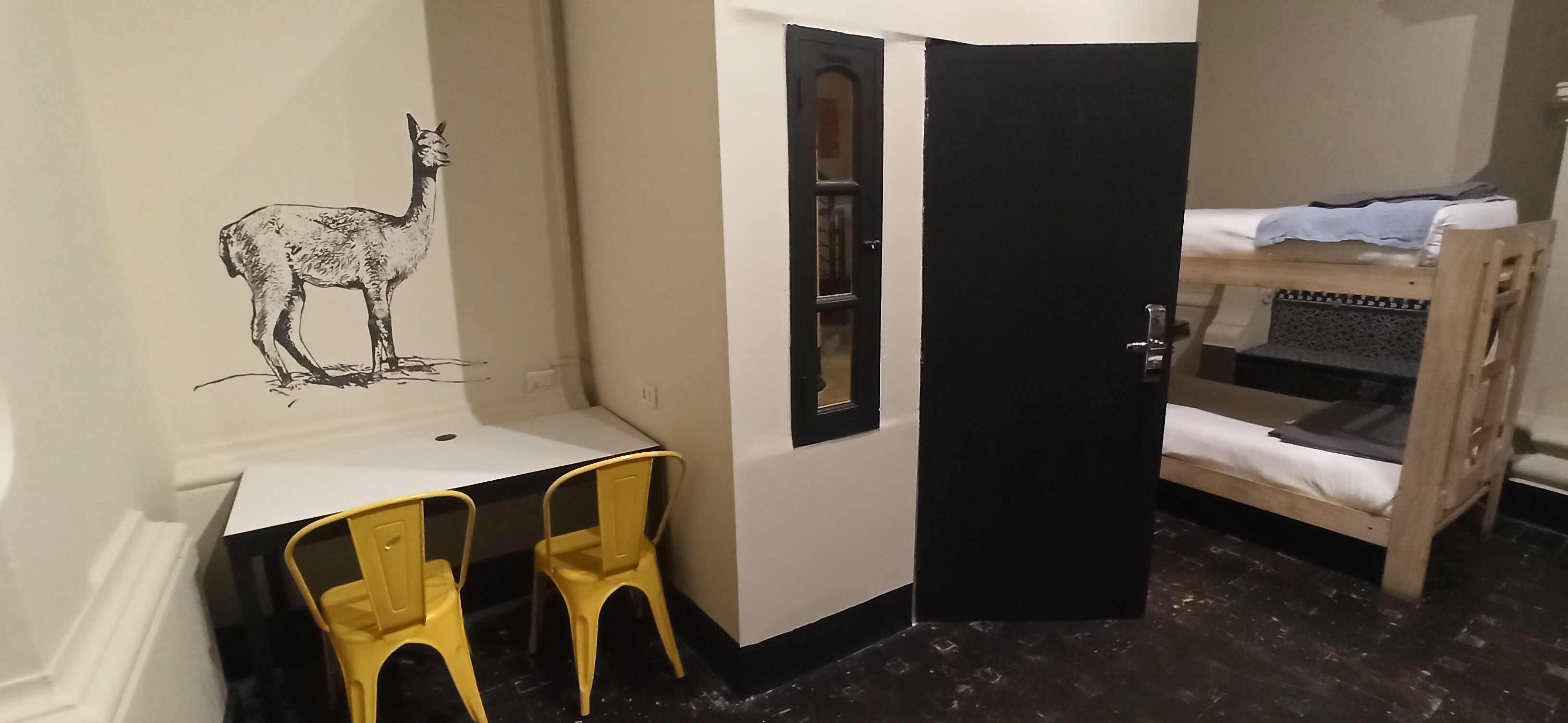Una habitación con un escritorio, dos sillas amarillas, una cama individual y un mural en la pared con un guanaco.