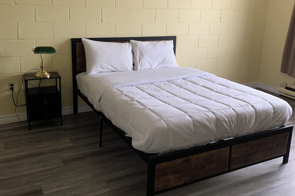 Una cama individual con estructura de madera y ropa de cama blanca en una habitación con pared de ladrillo y suelo de madera.