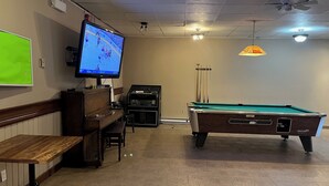 A billiards room with a pool table, a television displaying a sports game, a piano, and a green screen. - Spanish River Inn (Spanish)