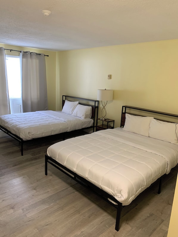Dos camas individuales con estructura de madera y ropa de cama blanca, una mesita de noche con lámpara y una ventana con cortinas.
