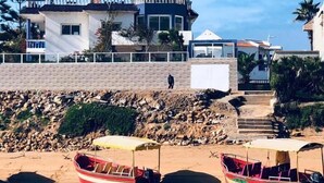 Escena costera con dos barcos amarrados en un muelle, un edificio con balcón y una costa rocosa.