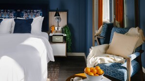 A modern hotel room with a bed, a chair, a bedside table with a lamp, a bowl of oranges, and a book.